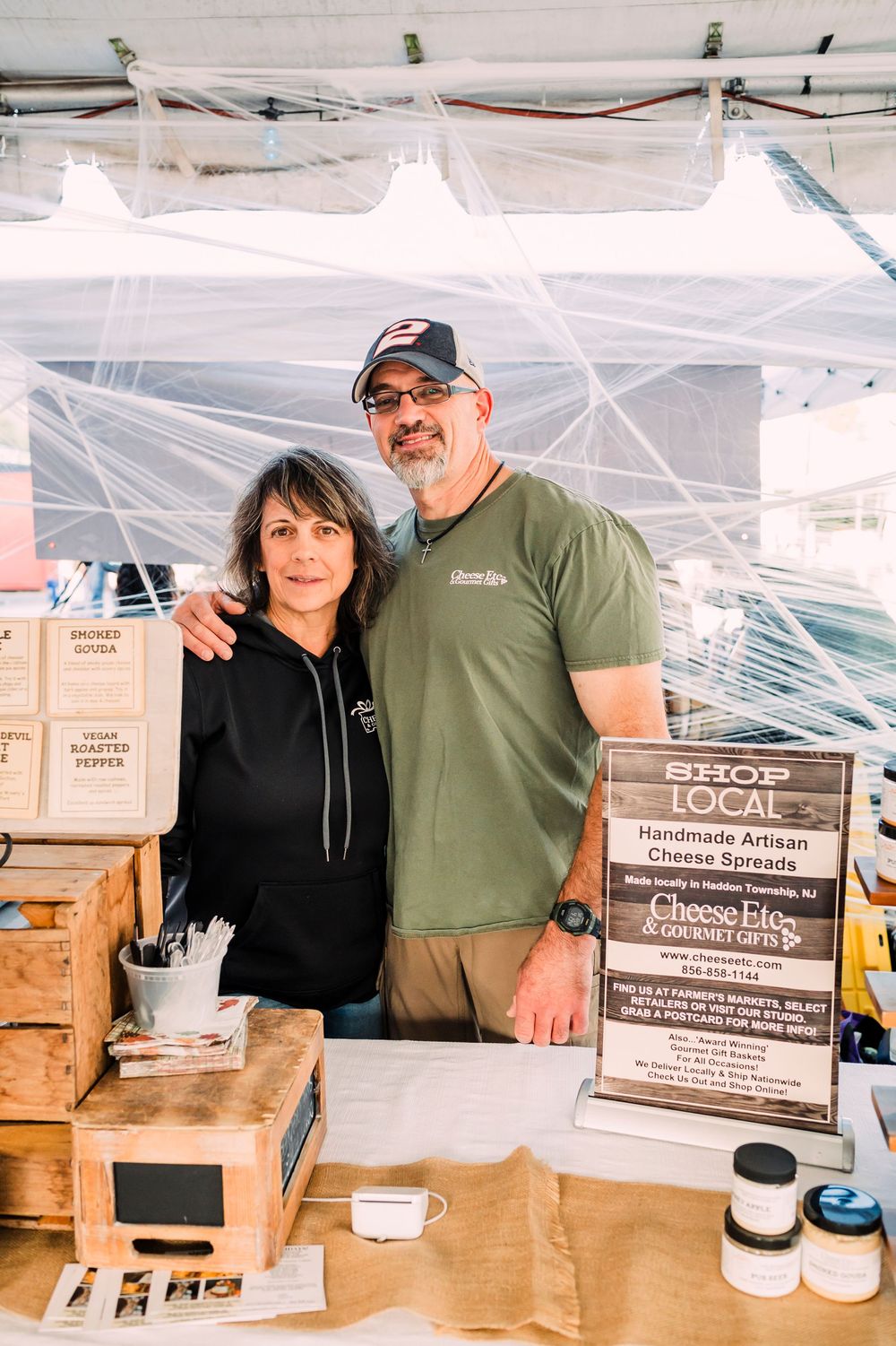 Vendors at a farmers market booth display local products and signage under a white tent canopy.