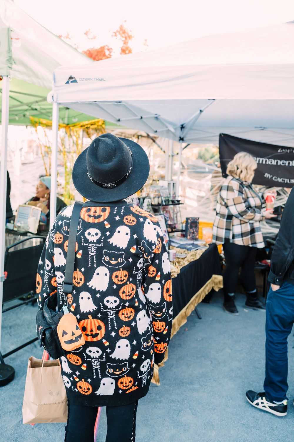 Someone wearing a Halloween-themed jacket with ghosts and pumpkins browses at an outdoor market.