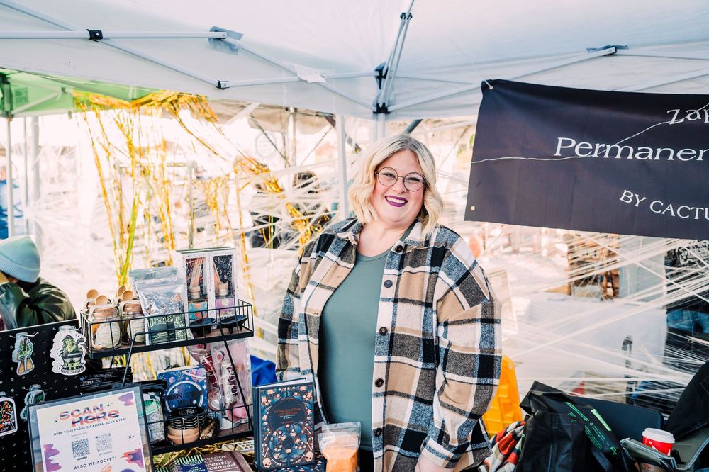 Vendor smiling at outdoor craft market booth displaying merchandise under white tent canopy.