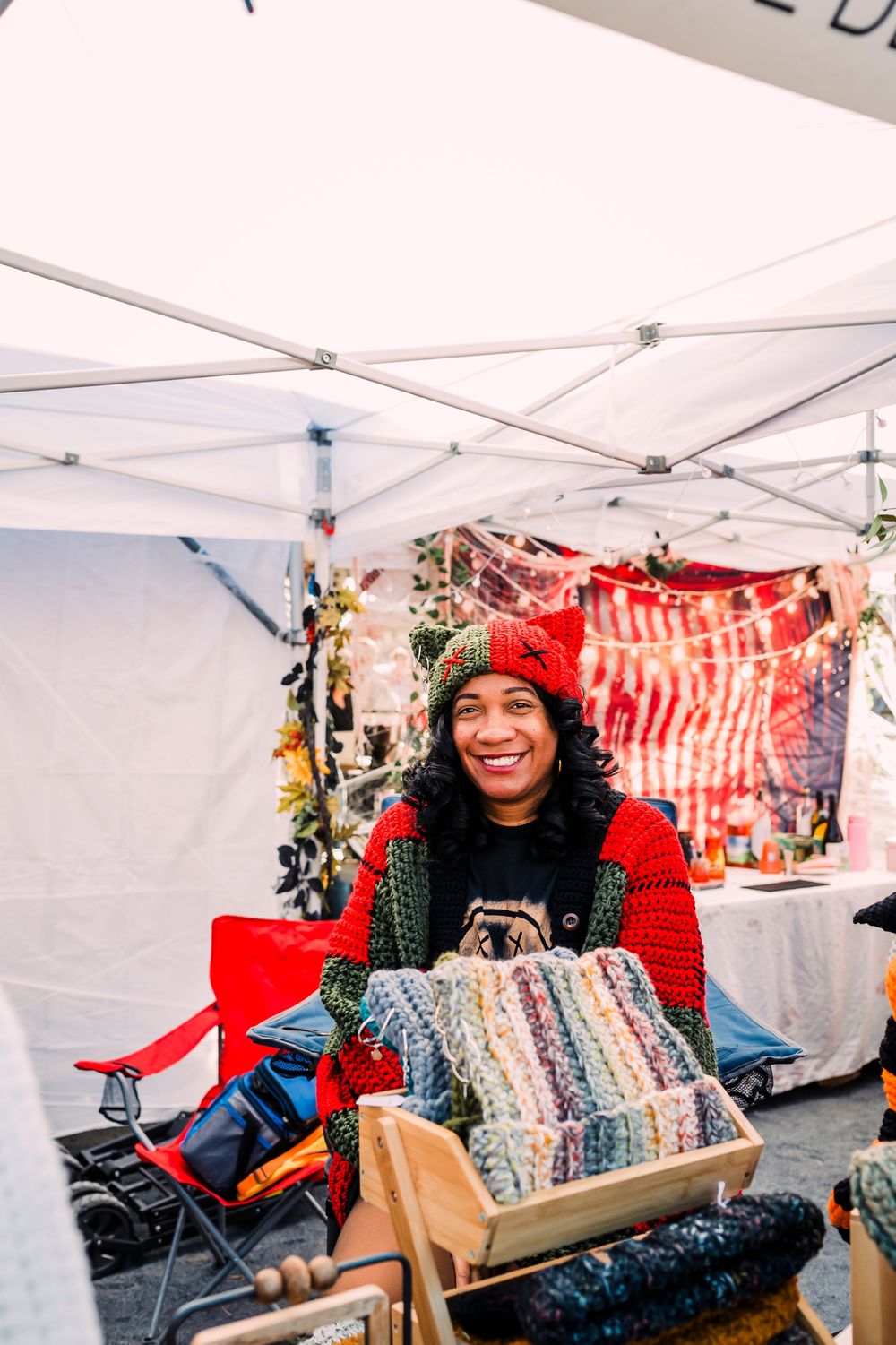 Vendors with colorful knitted goods and handcrafted items displayed at a market stall under white tents.