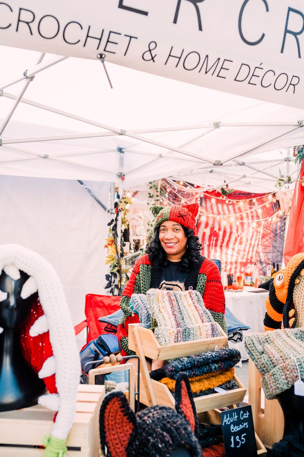 A craft vendor displays handmade crochet items at an outdoor market stall with colorful textiles and accessories.