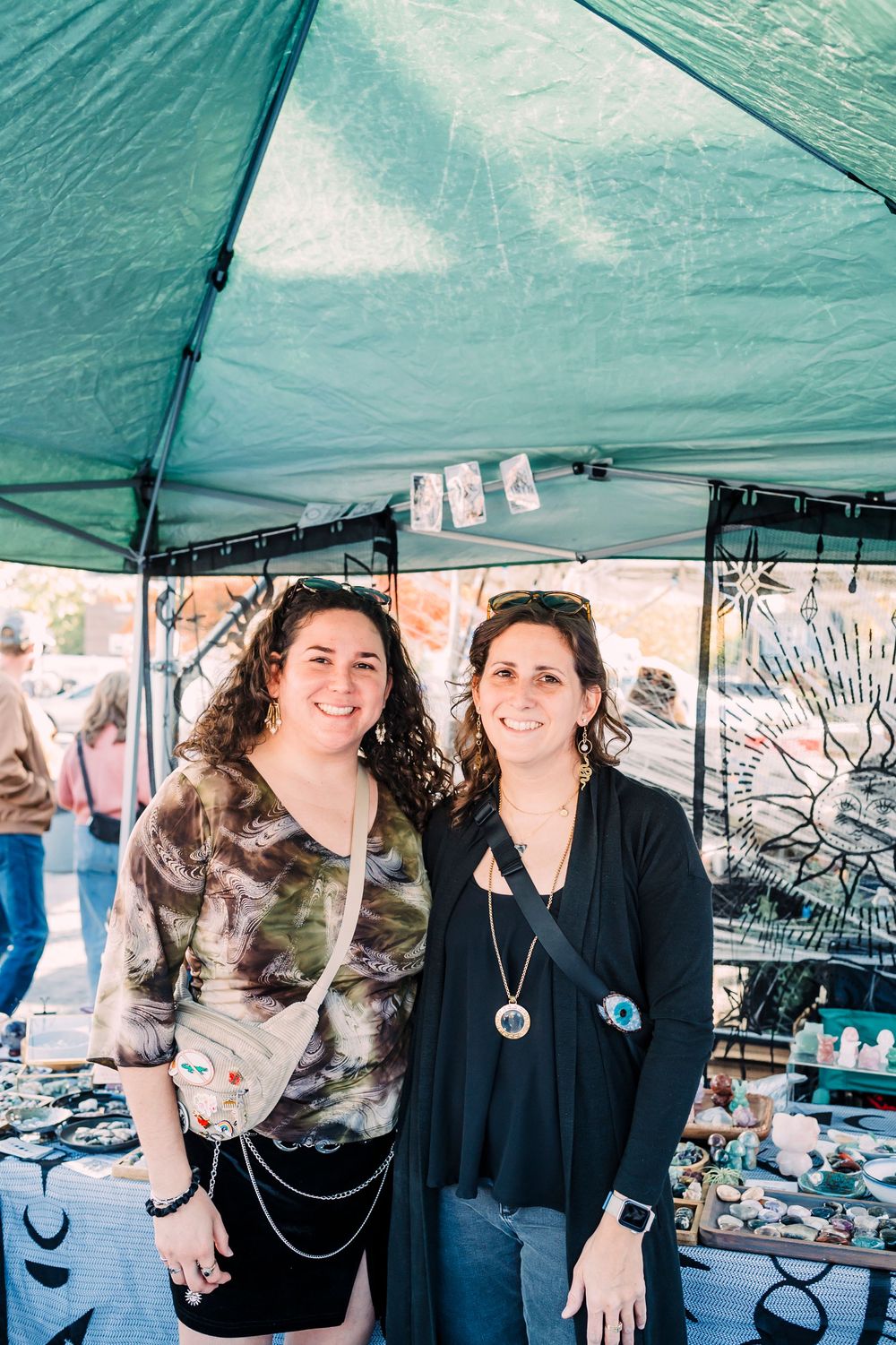 Two people stand together under a green canopy tent at what appears to be an outdoor market or craft fair.