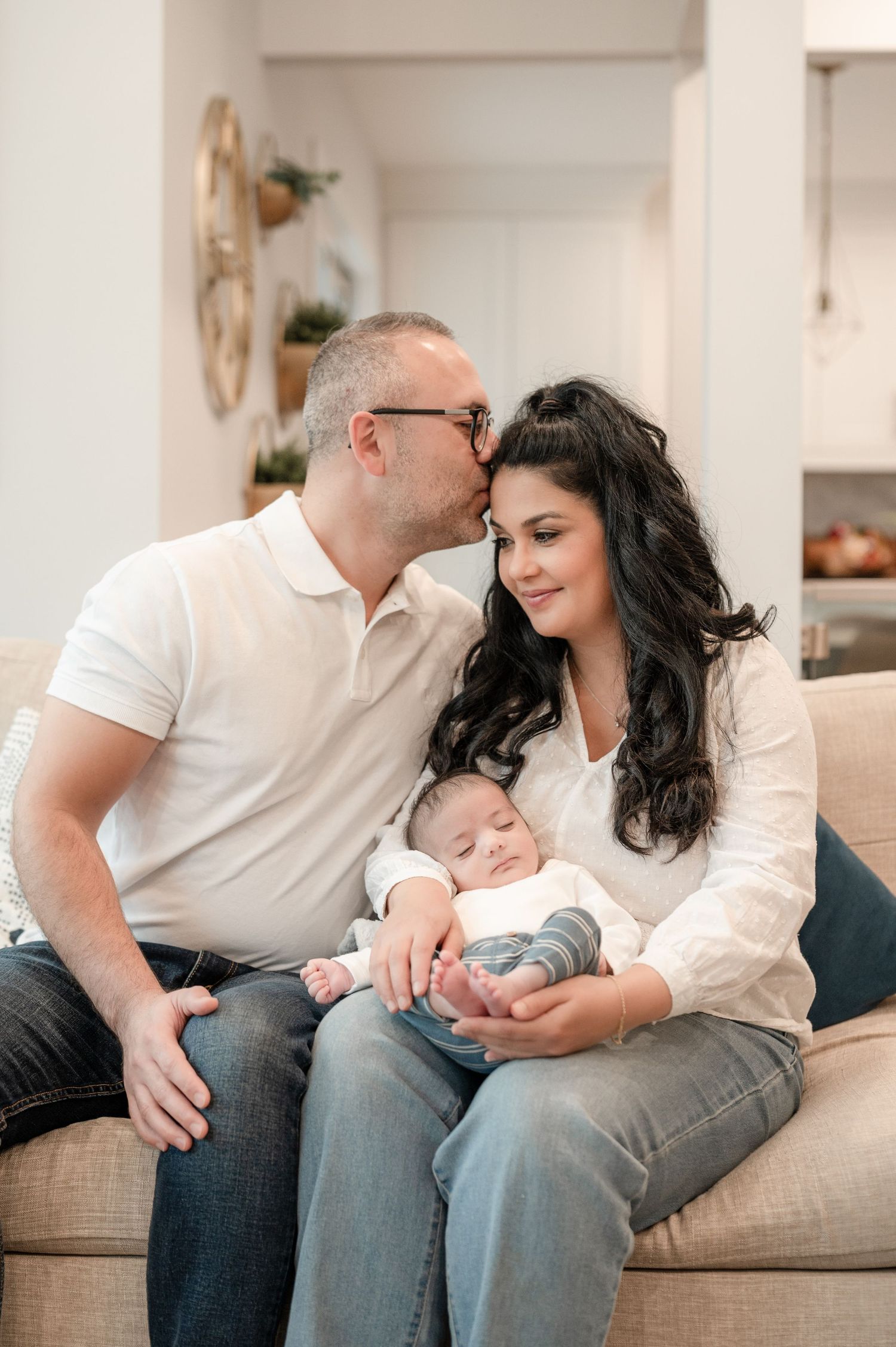 A family with a newborn rests together on a grey couch in a light-filled living room during an intimate photo session.
