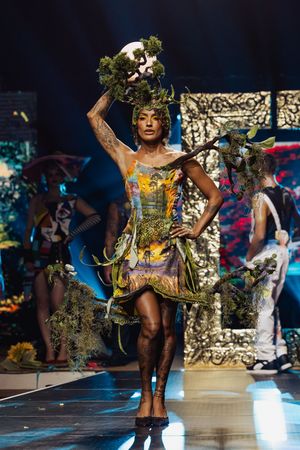Artistic fashion show performer in vibrant nature-inspired costume with moss and botanical elements on stage.