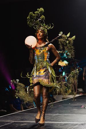 Fashion model strikes dynamic poses wearing an avant-garde green nature-themed costume on dark runway.