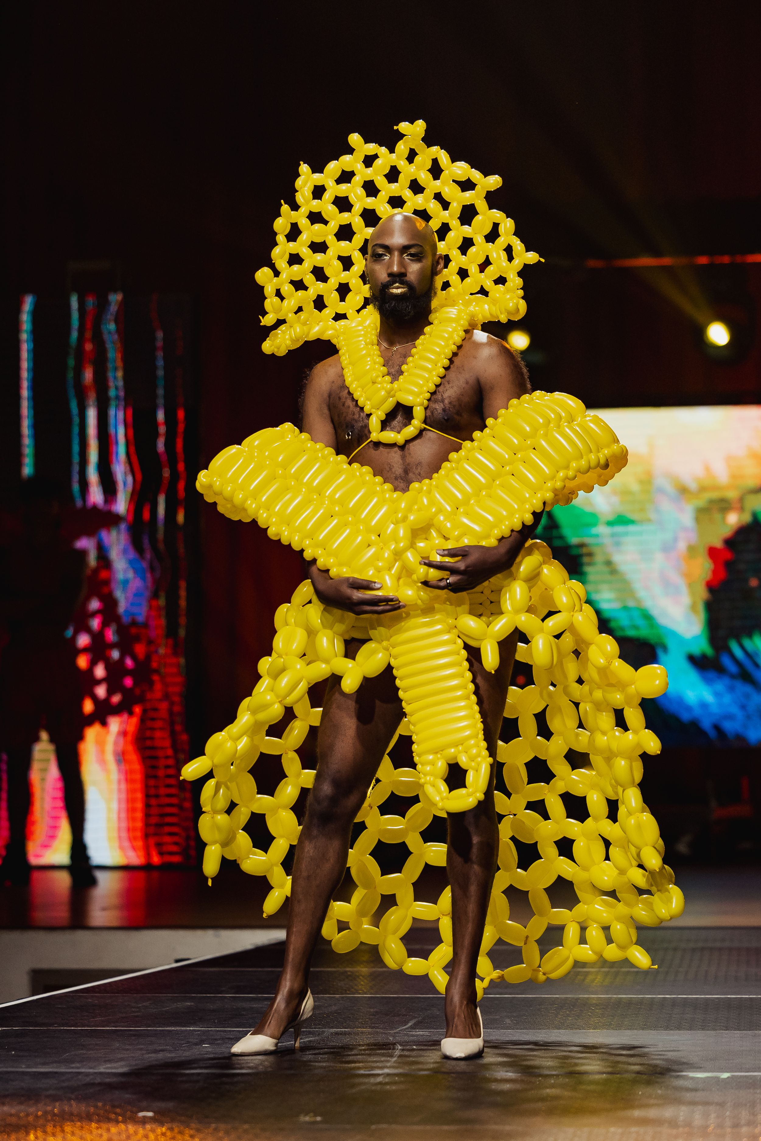 Performer wearing dramatic sculptural yellow balloon art costume dances on stage during fashion show performance.