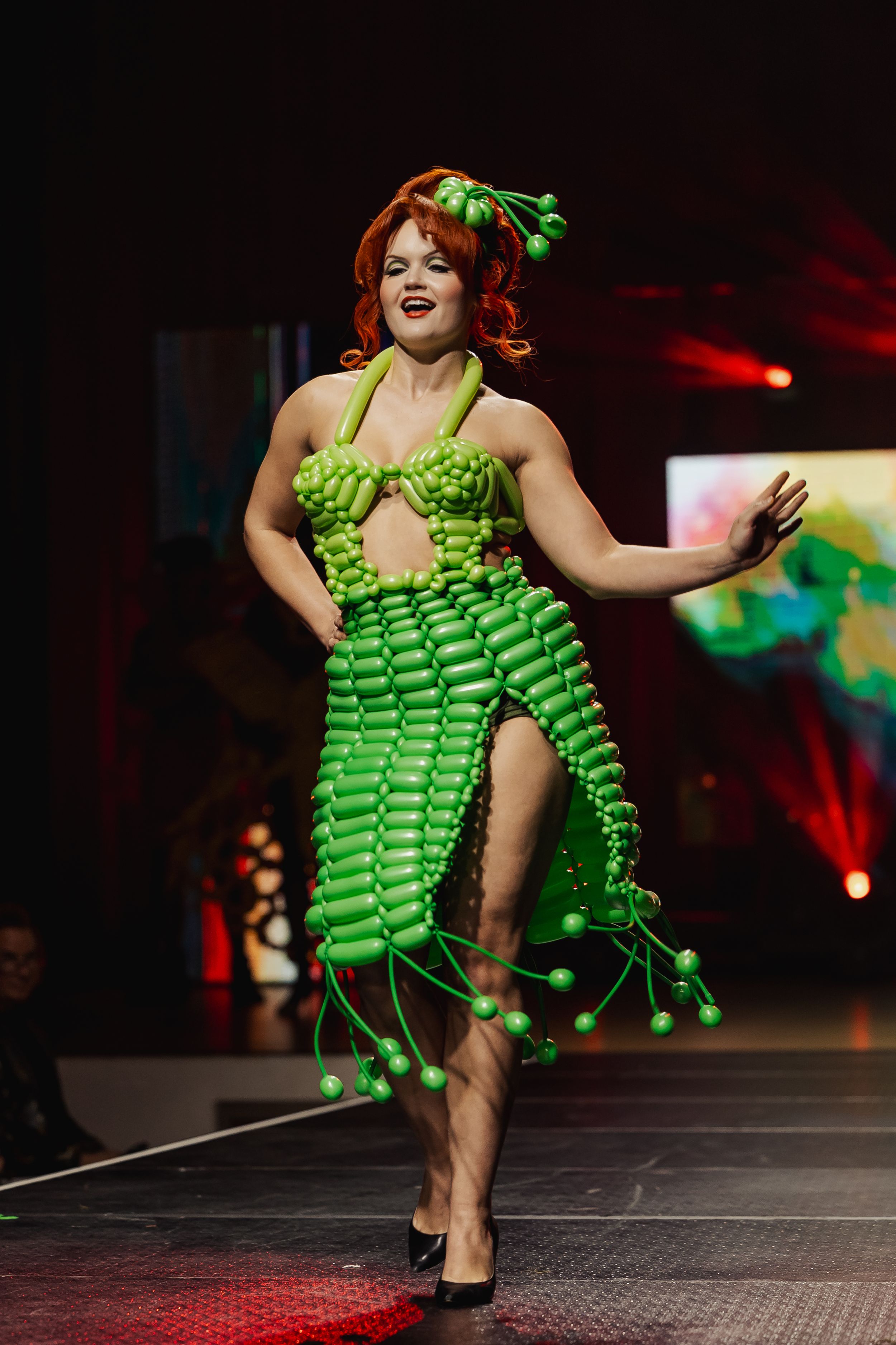 A performer in a unique lime green balloon dress strikes dynamic poses on a dark runway stage with dramatic lighting.