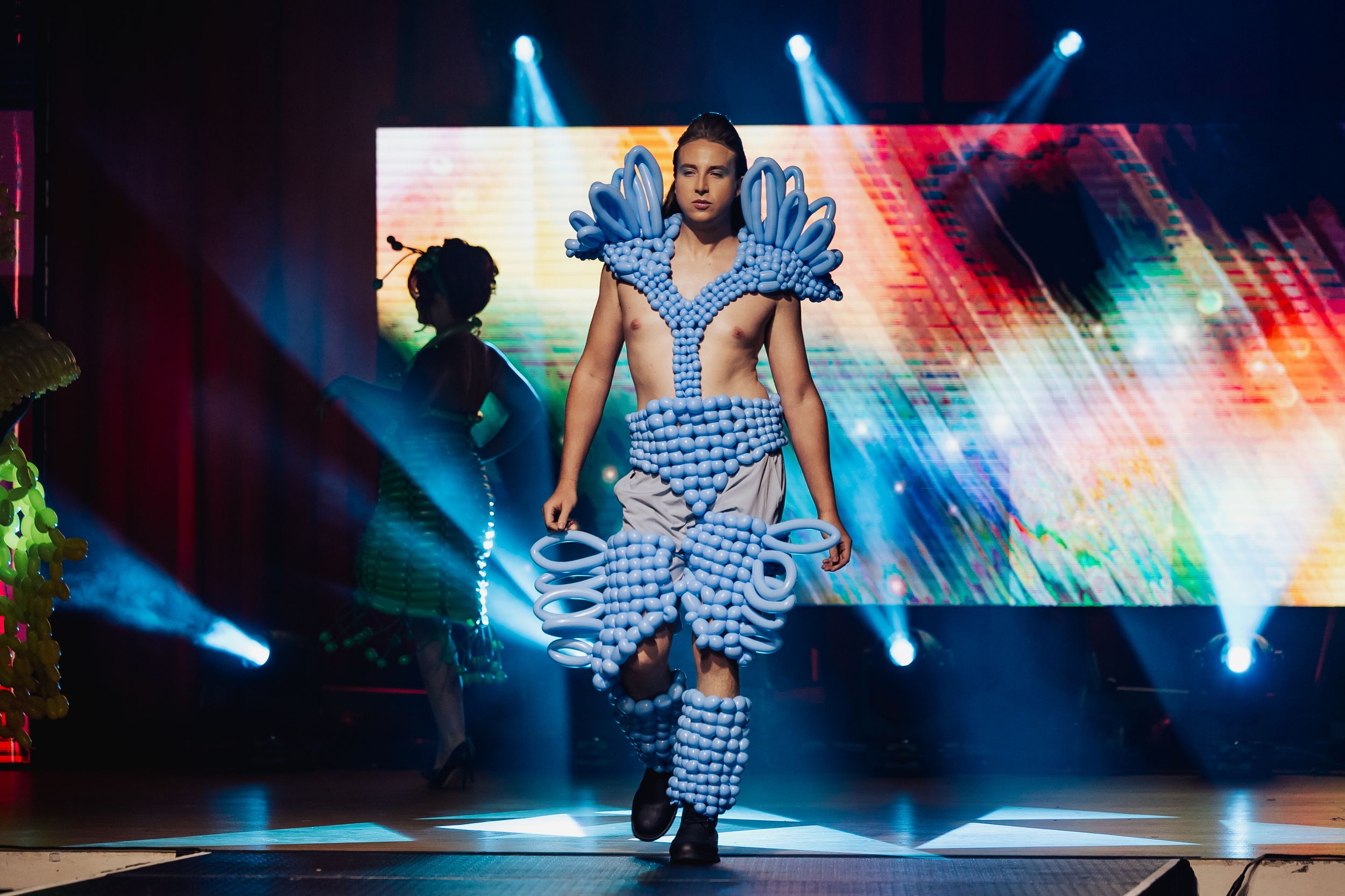 Dancer performing dynamic moves on illuminated stage with colorful lights and dramatic blue spotlights in background.