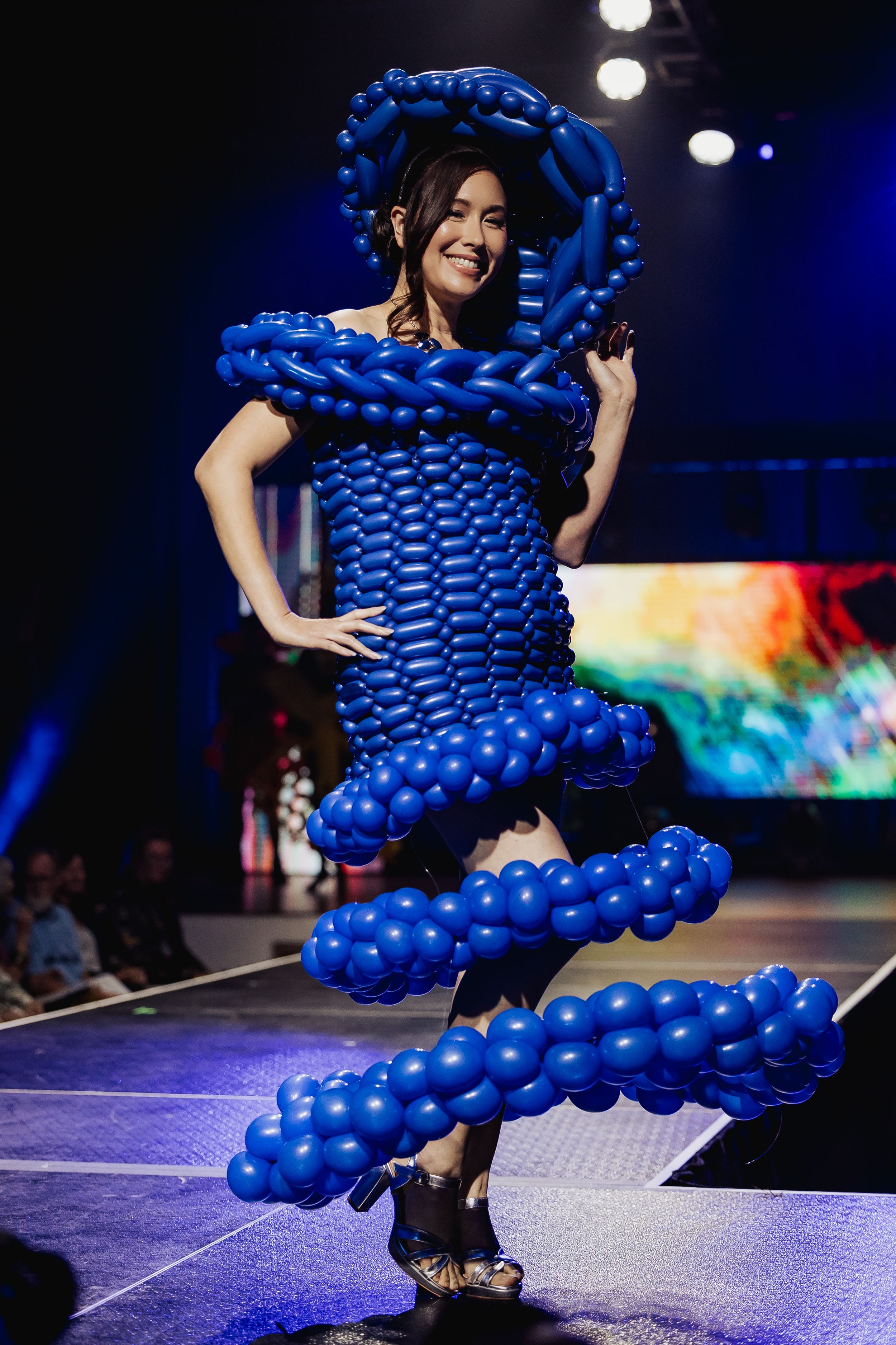 A performer wears a dramatic dress made entirely of blue balloons on a darkened stage with colorful lighting effects.