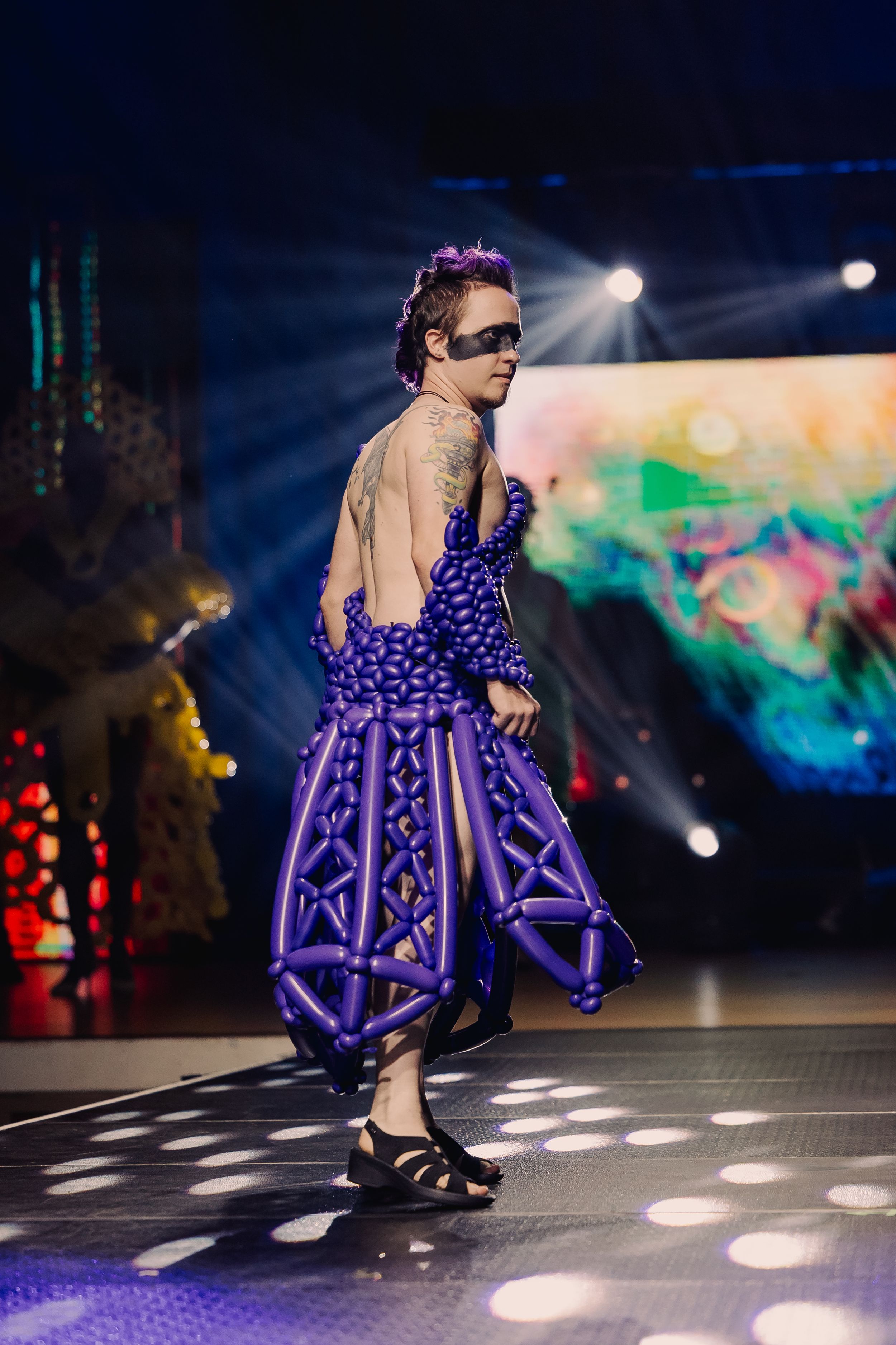 Fashion model walks runway wearing dramatic purple tiered dress with structural ruffles under dramatic evening lighting.