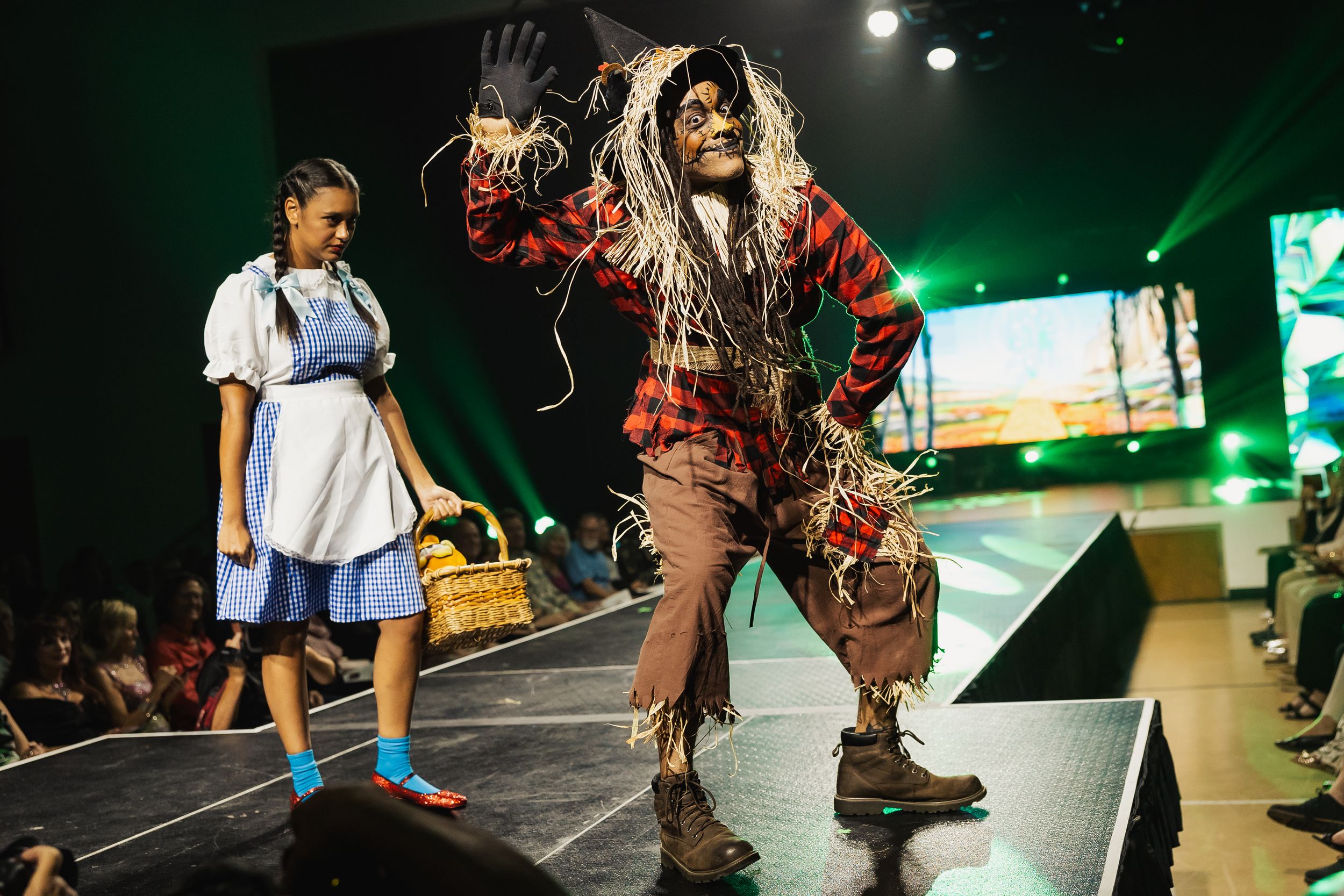 Performers in floral costumes and boots dance energetically on a dark stage with dramatic green lighting effects.