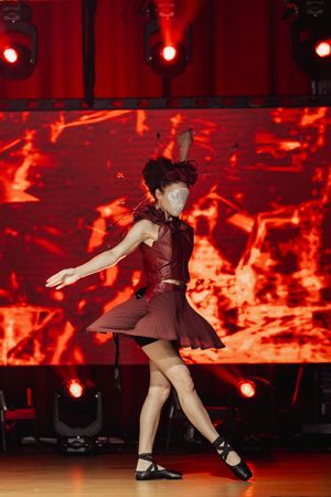 Dancer performs dynamic movements against dramatic red lighting and stage backdrop with fiery visual effects.