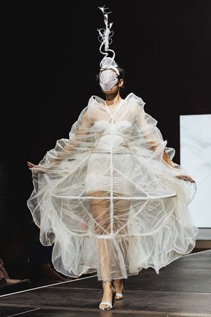 Fashion model walks runway in dramatic white ruffled translucent dress with voluminous layered design on dark stage.