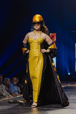 Model walks runway wearing bright yellow textured jumpsuit with black flowing cape at fashion show against blue backdrop.