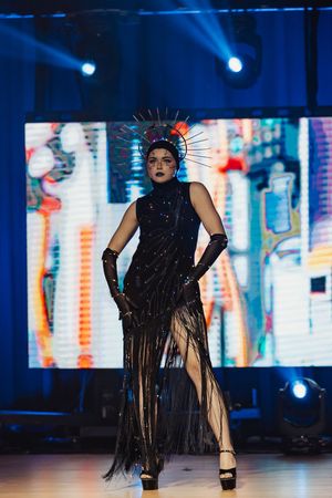 Model walks runway in dramatic black fringe dress with platform shoes during fashion show with blue lighting.