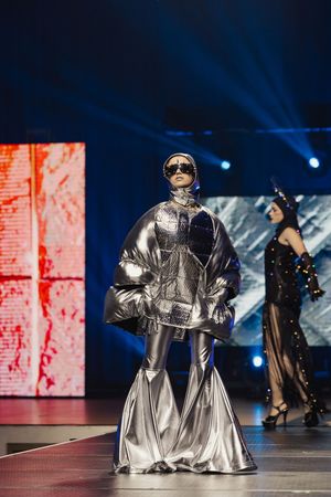 Models showcase futuristic silver metallic gowns with dramatic silhouettes on a dark runway with blue lighting effects.