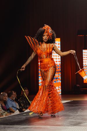 A performer in a striking orange ruffled dress twirls on stage with dramatic lighting in the background.
