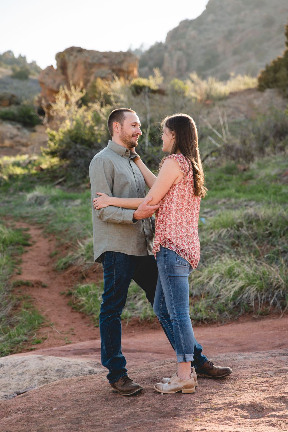 A couple shares an intimate moment together on a red dirt trail surrounded by desert landscape at golden hour.