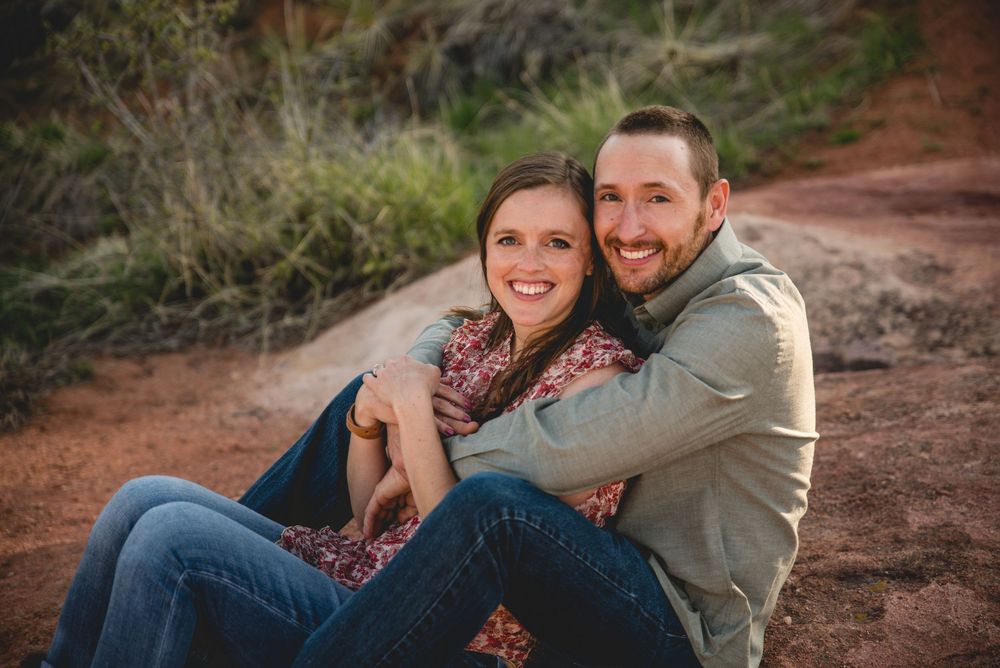 A couple sits together laughing on a red dirt path during an outdoor photo session.