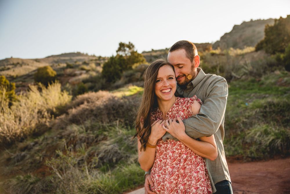 A romantic couple embraces and laughs together on a hiking trail during golden hour with hills in the background.