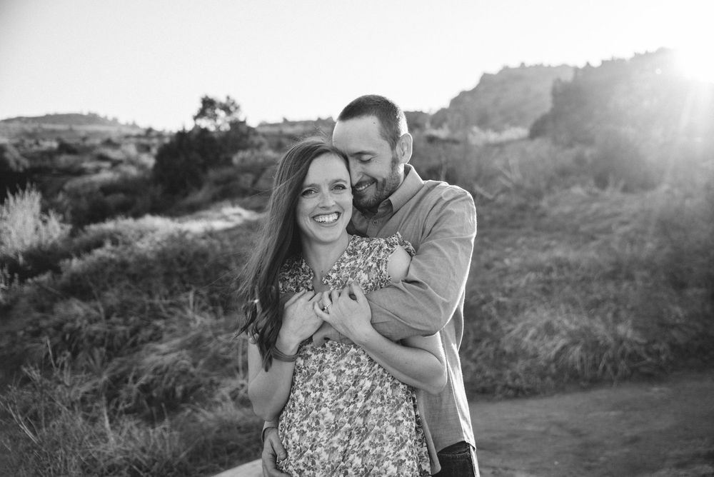 A black and white portrait of a couple embracing and smiling on a hilltop at sunset.
