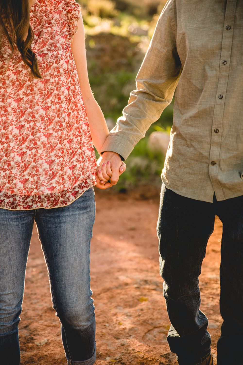 A couple holds hands while standing on a dirt path, wearing a floral dress and beige button-down shirt with jeans.