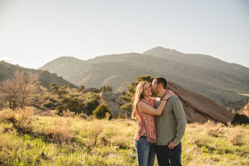 Couple embraces while standing in golden mountainside field during sunset with California hills in background.