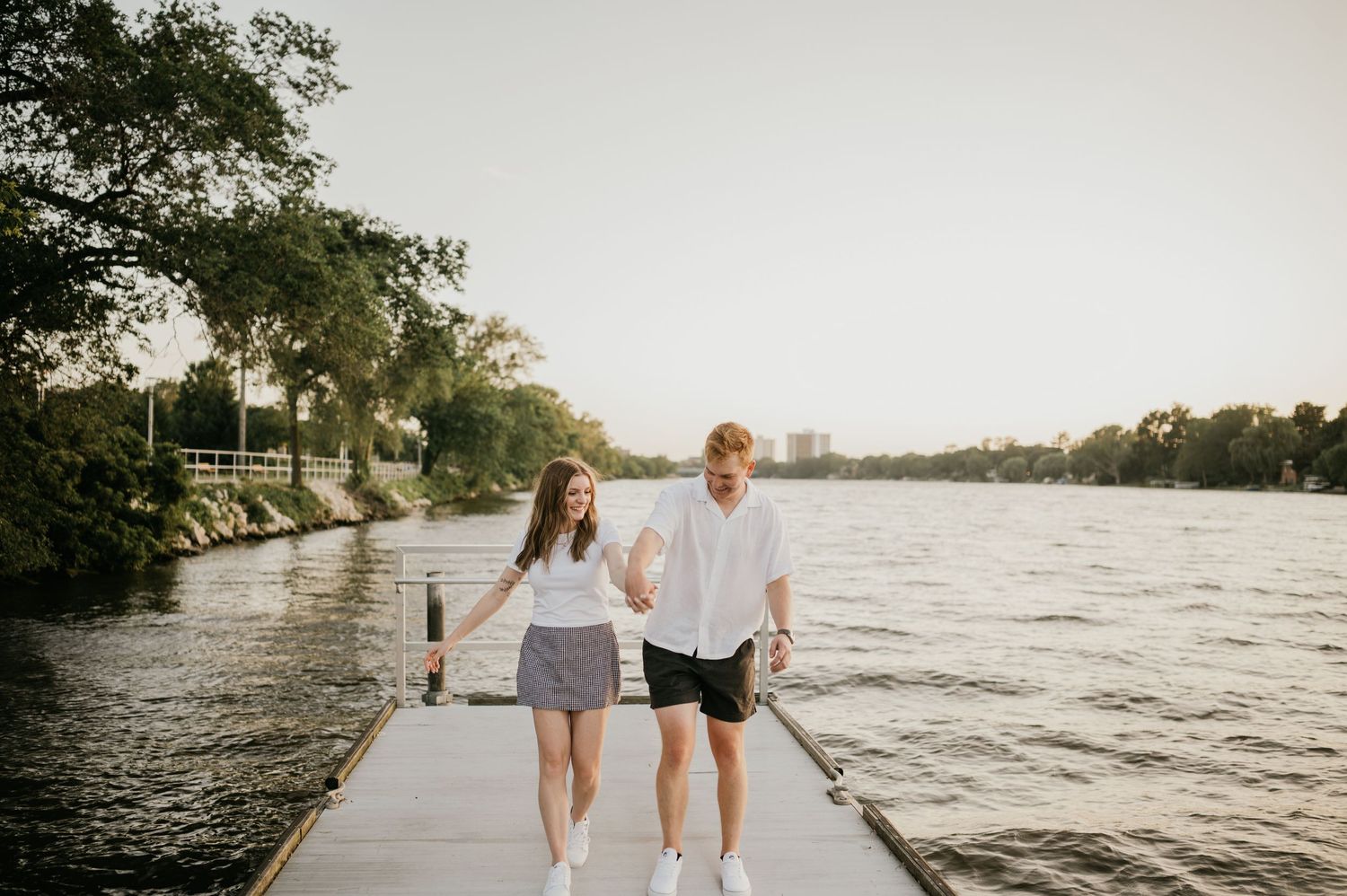Two people holding hands while walking on a pier by a calm river during sunset.