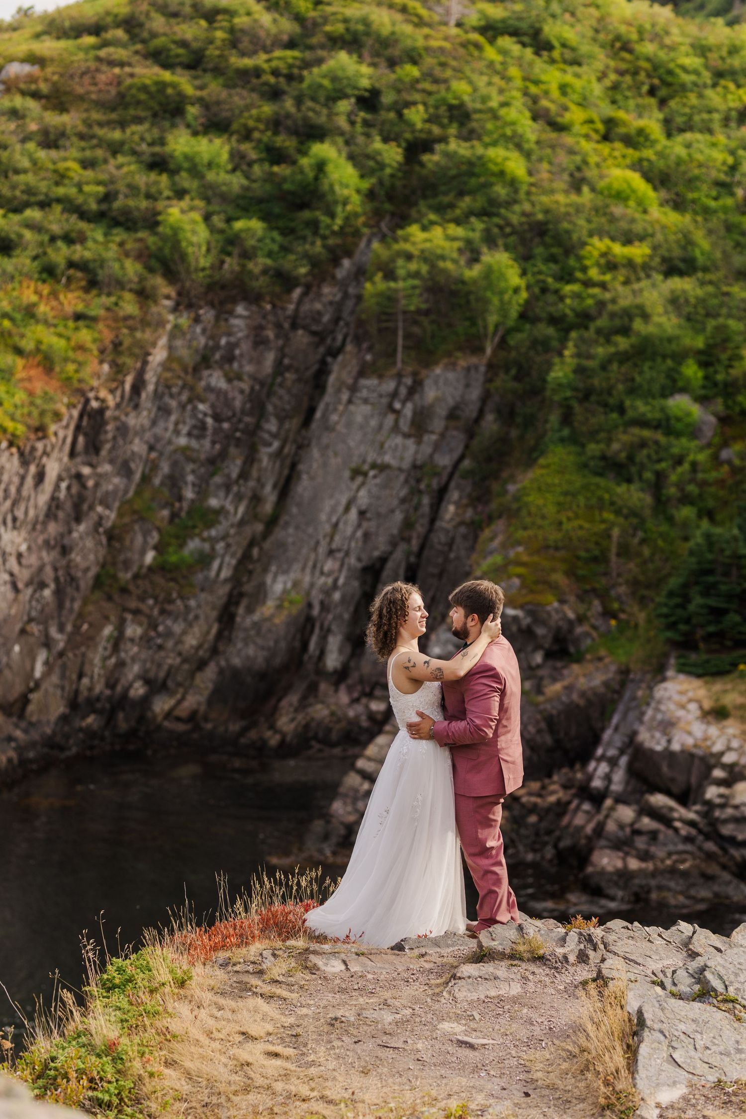 A couple embraces on a rocky cliff edge against a dramatic backdrop of tall cliff walls and lush greenery.