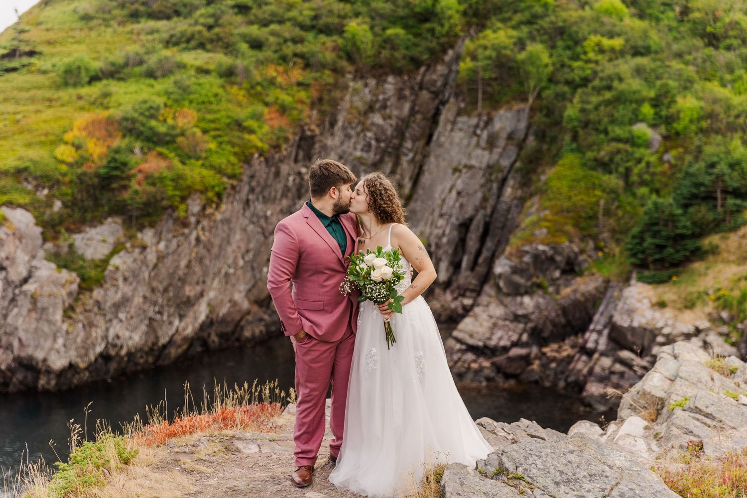 A couple in wedding attire shares an intimate moment on a rocky cliff overlooking scenic mountain landscapes.