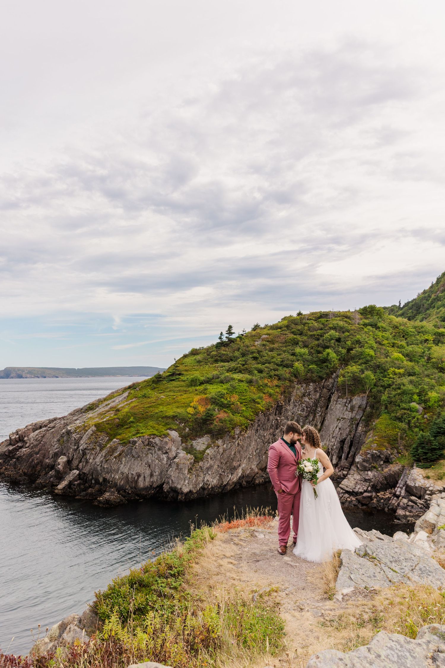 A scenic coastal wedding ceremony takes place on a rugged cliff overlooking the ocean against cloudy skies.