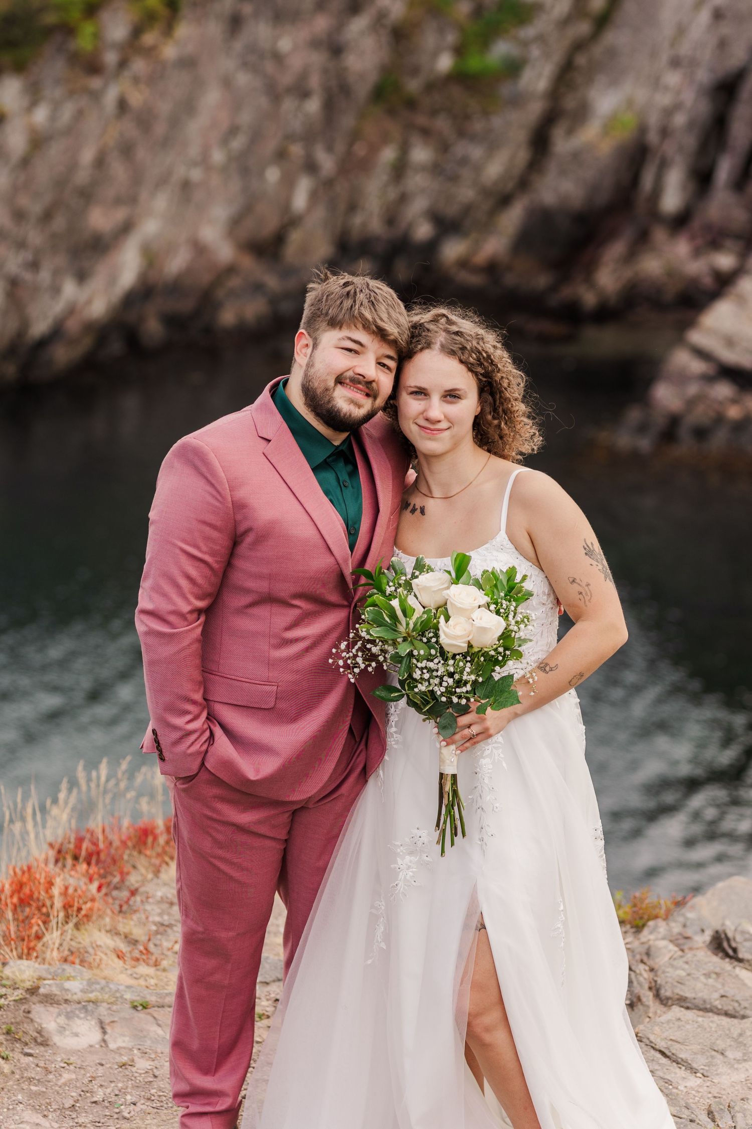 Couple in pink suit and white dress embrace near rocky cliffs and water during autumn wedding photoshoot.