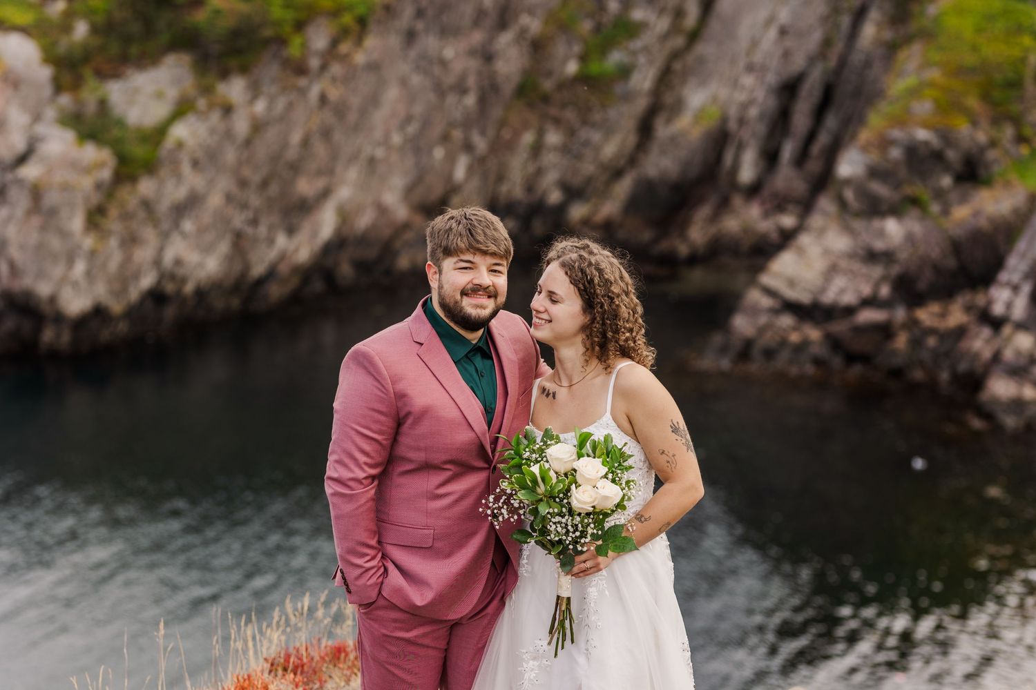 A happy newlywed couple shares an intimate moment against a backdrop of rocky cliffs and water.
