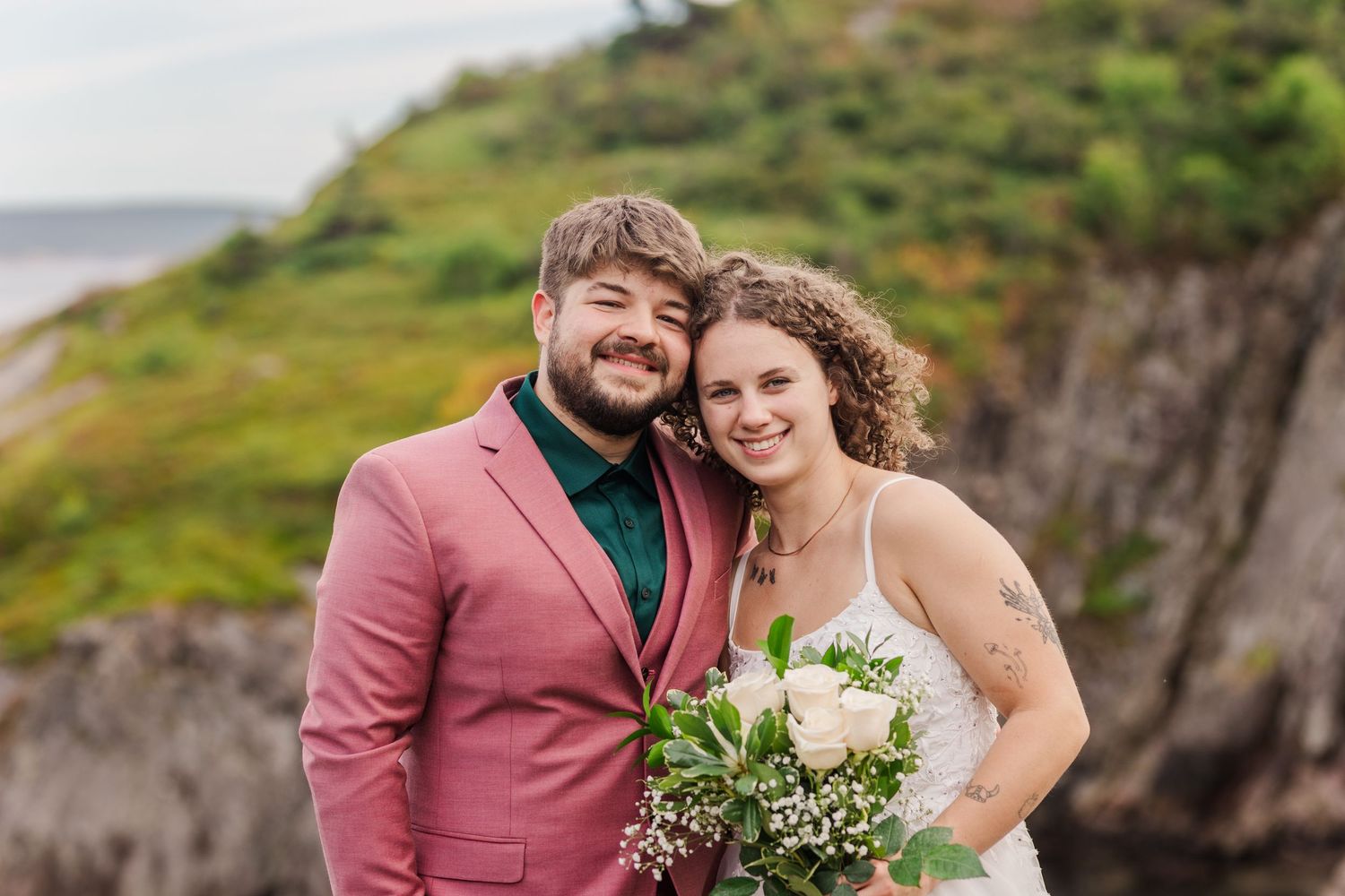 Wedding couple poses together on scenic coastal cliffs with green hills and ocean views.