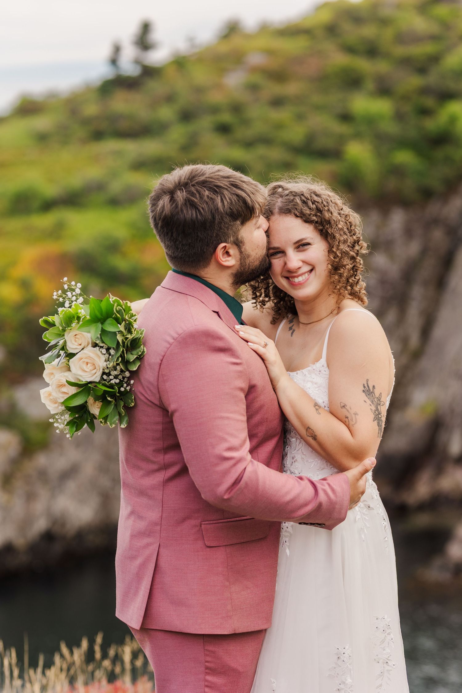 A romantic wedding couple embraces on a scenic cliffside overlooking water, with the bride in white and groom in pink.