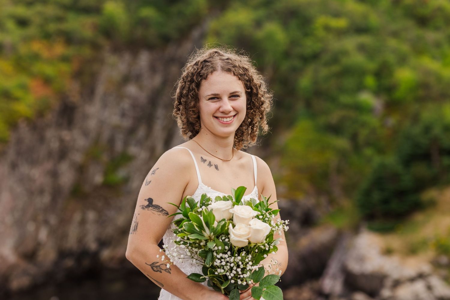 Bride in lace off-shoulder wedding dress holds white and green bouquet in rustic outdoor setting.