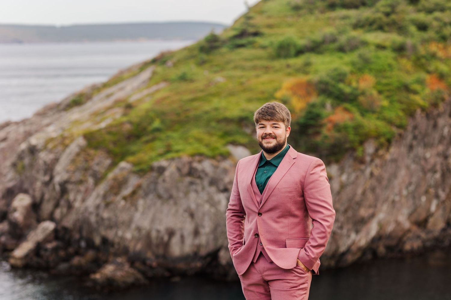 Couple embraces on dramatic coastal clifftop with stormy gray ocean and green landscape in background.
