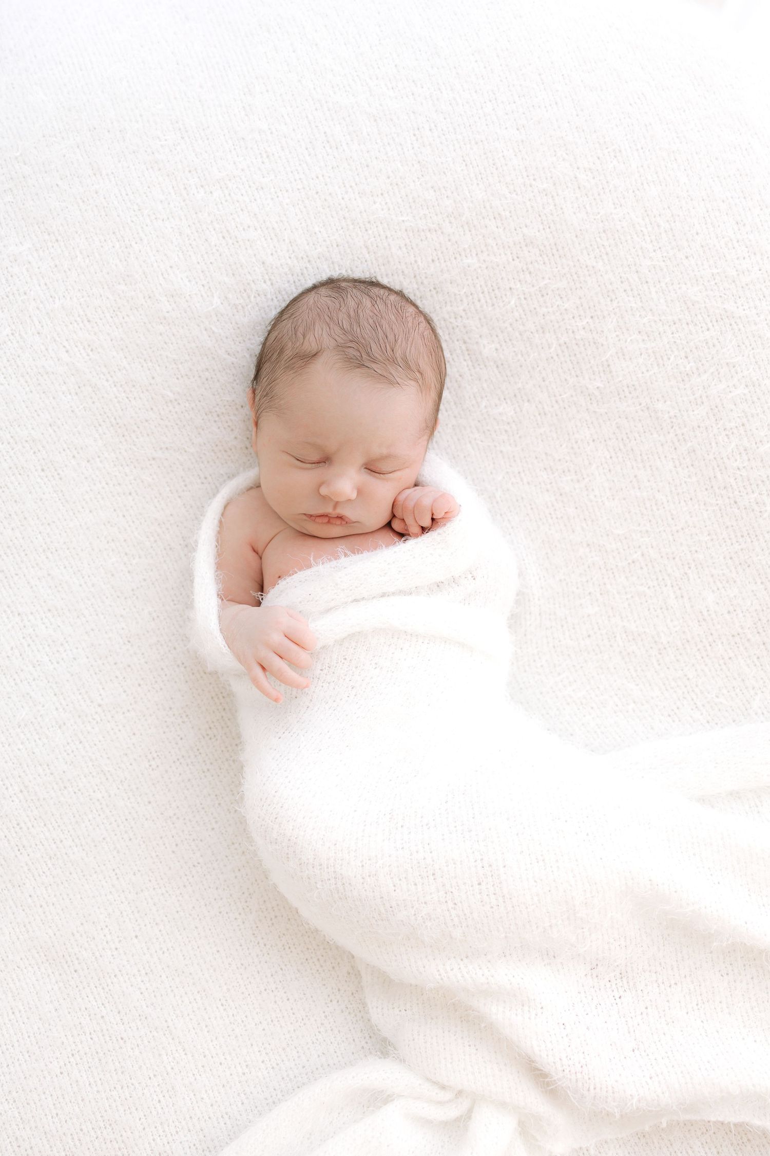 A tiny newborn baby wearing a white onesie lying peacefully on soft white bedding.