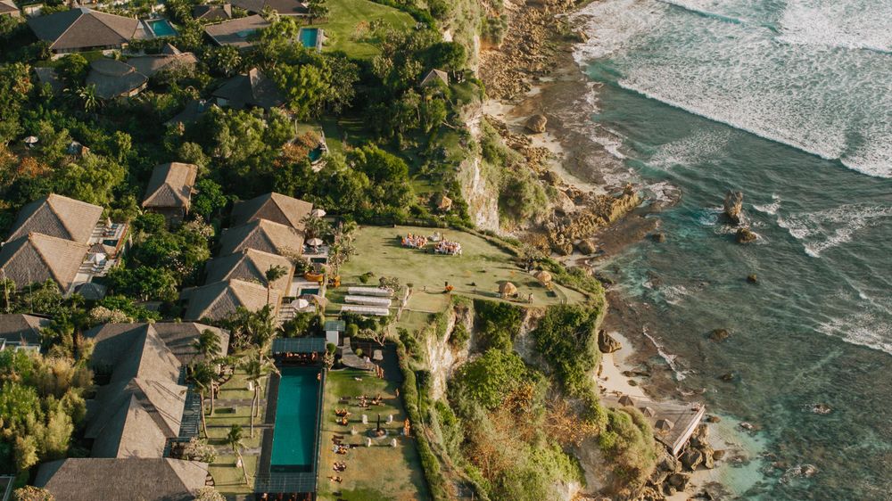 Aerial view of luxury cliffside villas with swimming pools overlooking the turquoise ocean waves in Bali.