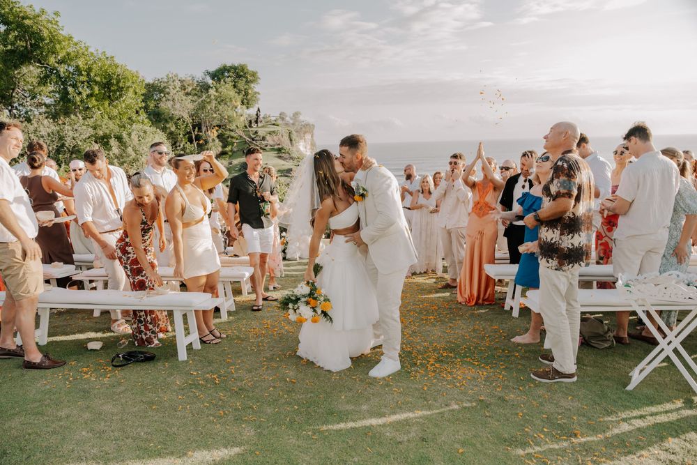 Wedding guests gather outdoors on a grassy lawn for a seaside ceremony during golden hour.