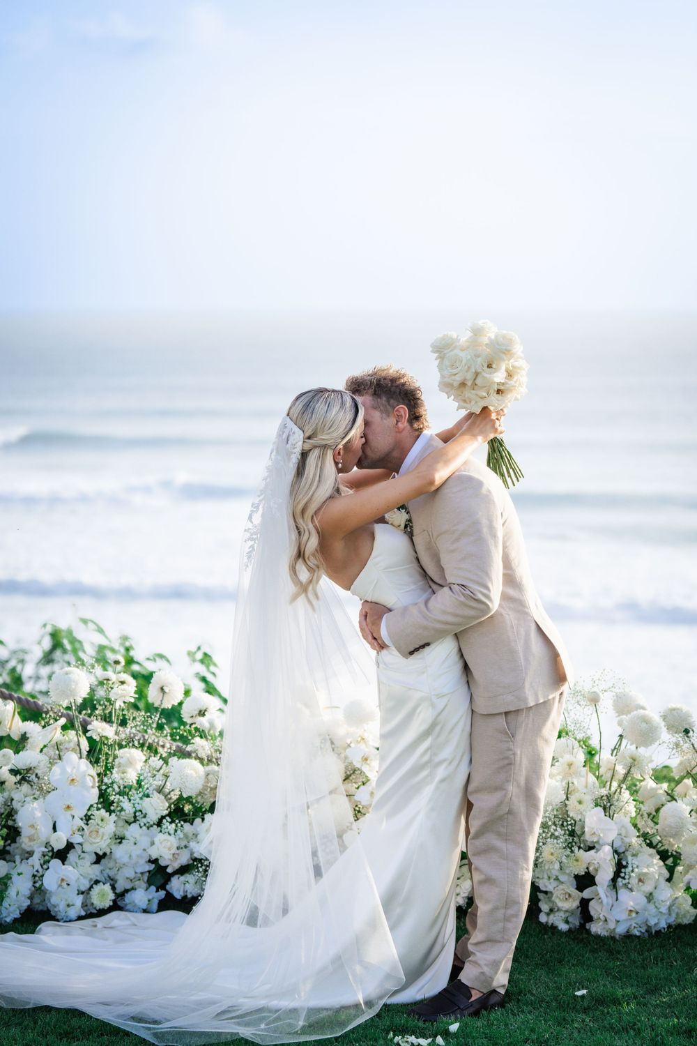 A romantic oceanside wedding with a couple embracing amidst beautiful white floral arrangements and a flowing veil.