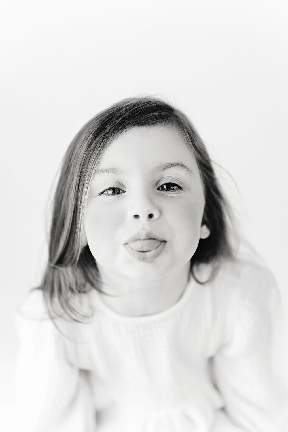 Black and white portrait of a young child making a playful kissing face while wearing a light colored top.