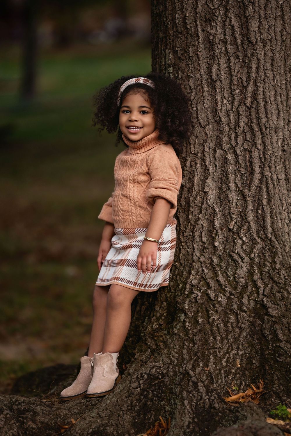 A small child in a peach sweater and plaid skirt sits against a large tree trunk in an autumn outdoor setting.