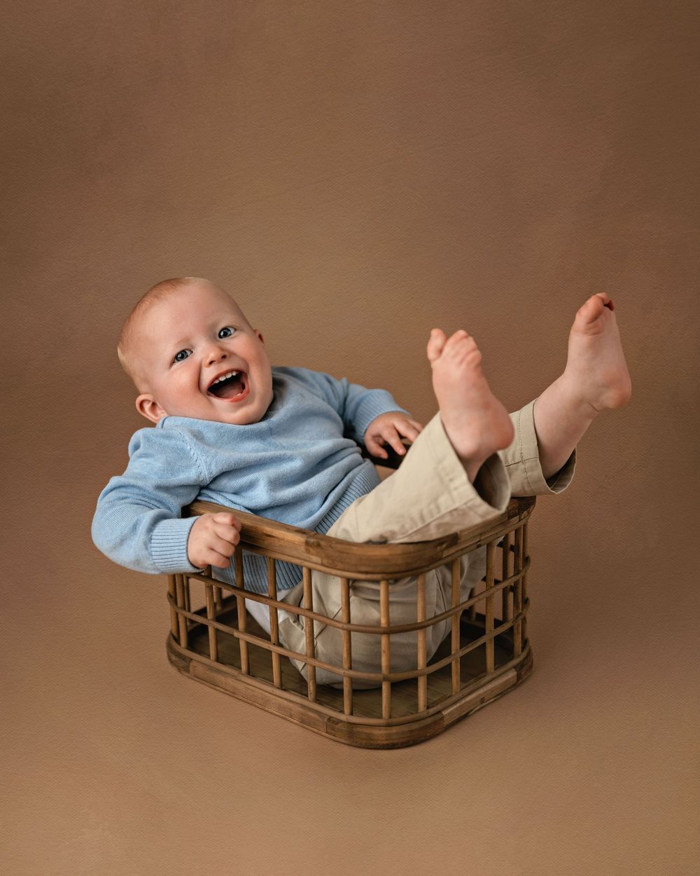 A baby in light blue clothing reclines in a vintage wooden basket against a warm brown studio backdrop.
