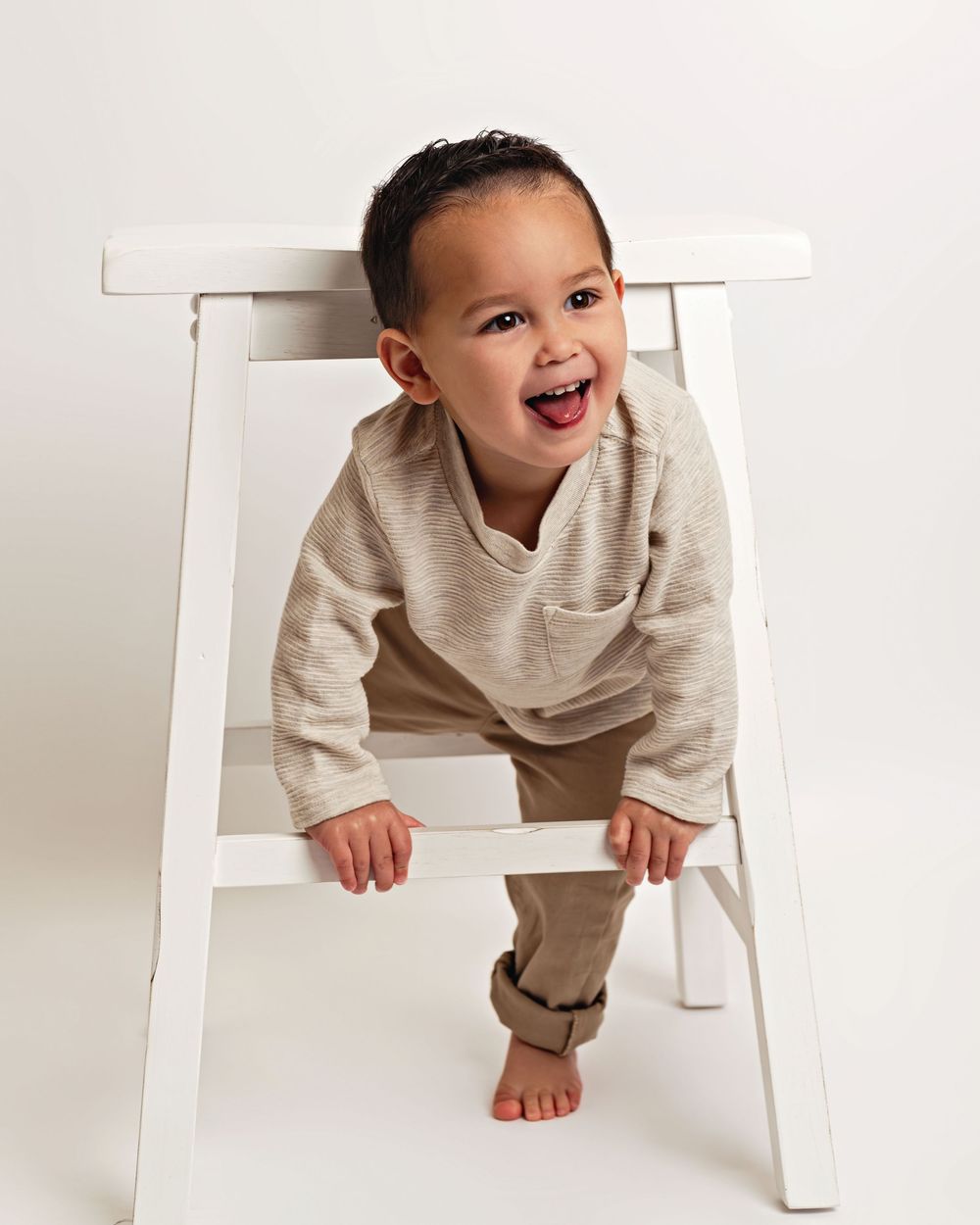 Child in beige outfit playing and climbing on white bench.