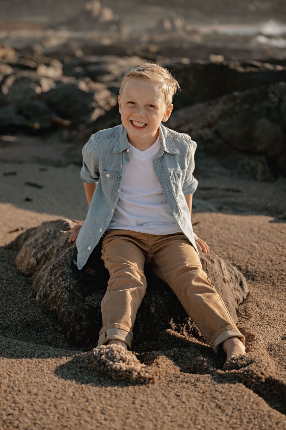 Child in denim jacket sitting on sandy beach during golden hour.