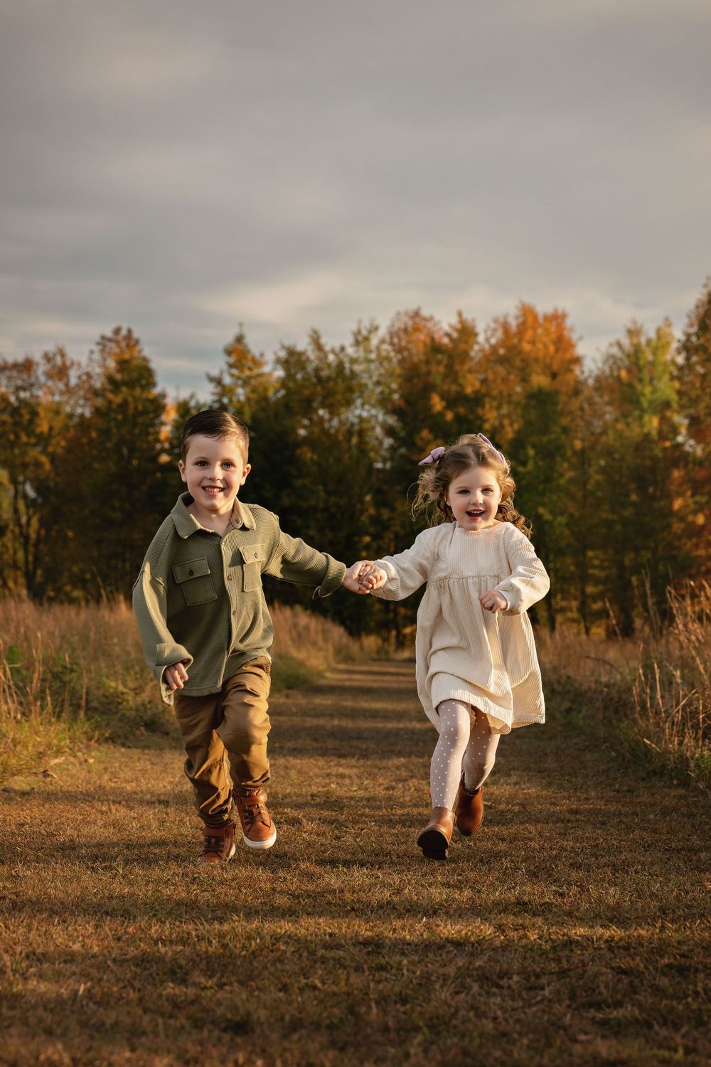 Two children holding hands while running together on a dirt path during autumn with fall foliage in the background.