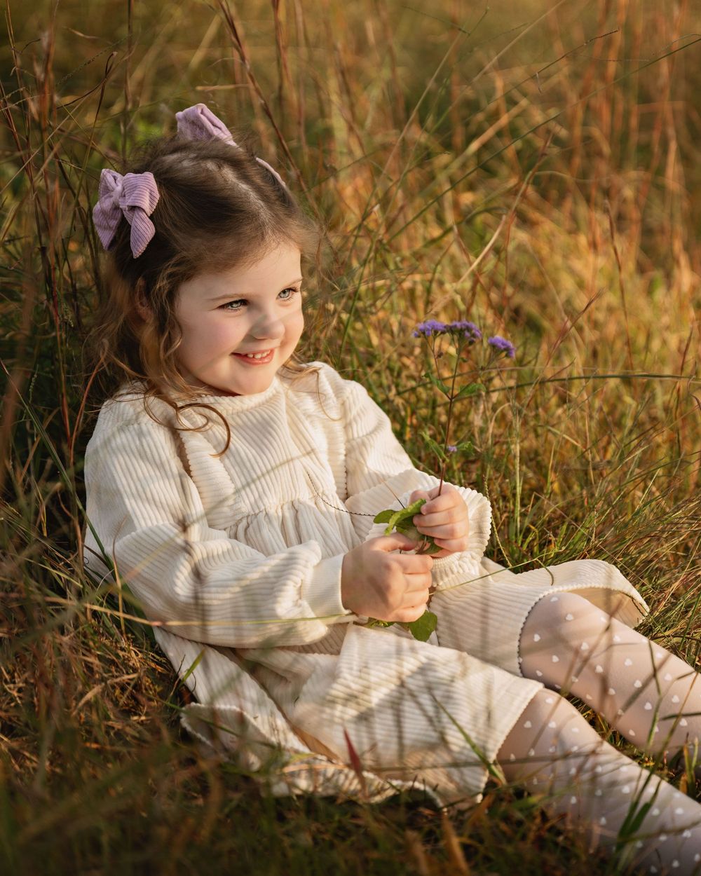 A child in a cream-colored dress sits in a meadow of tall grass and wildflowers during golden hour.