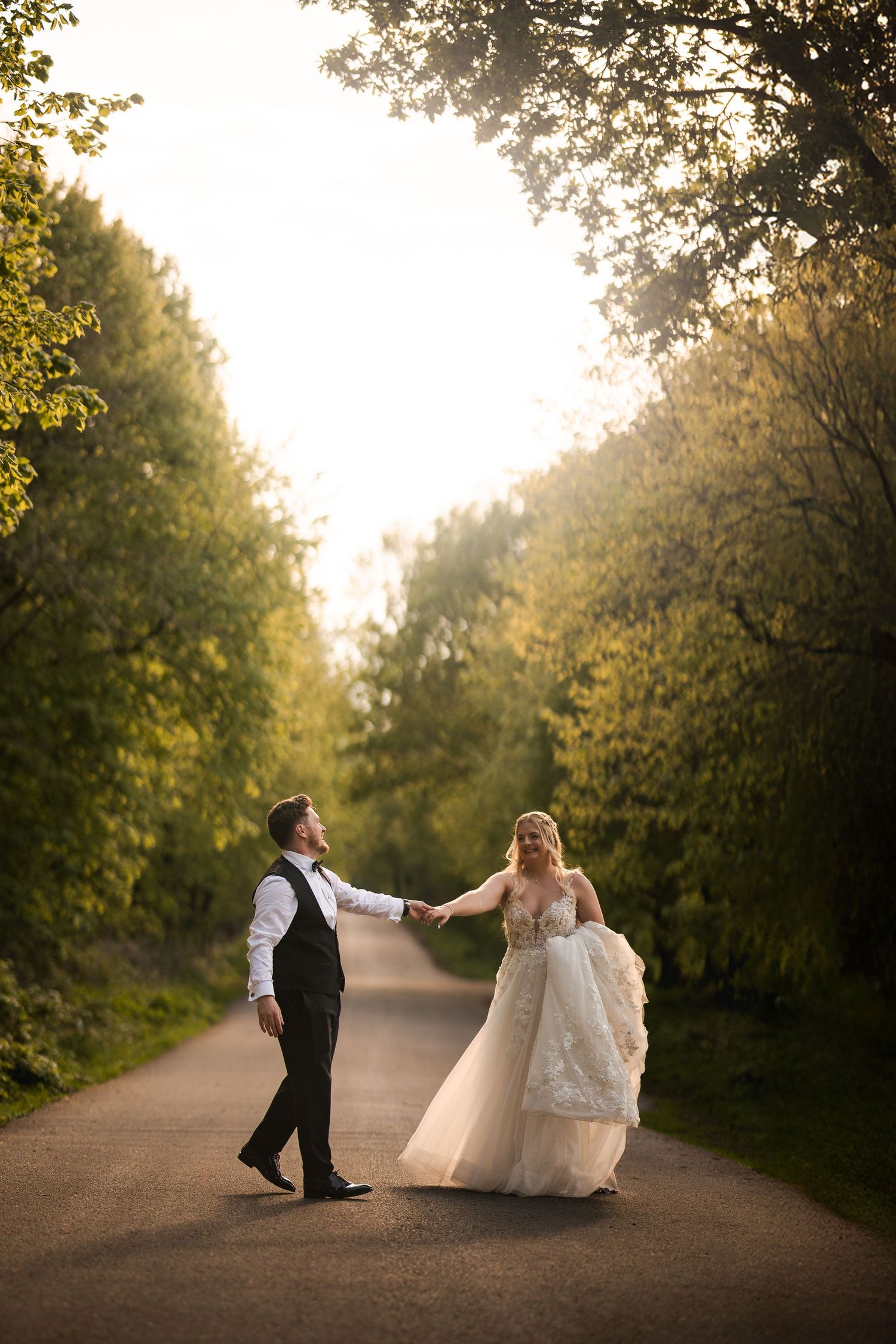 Couple in wedding attire dancing and twirling together on a scenic dirt path surrounded by trees at sunset.