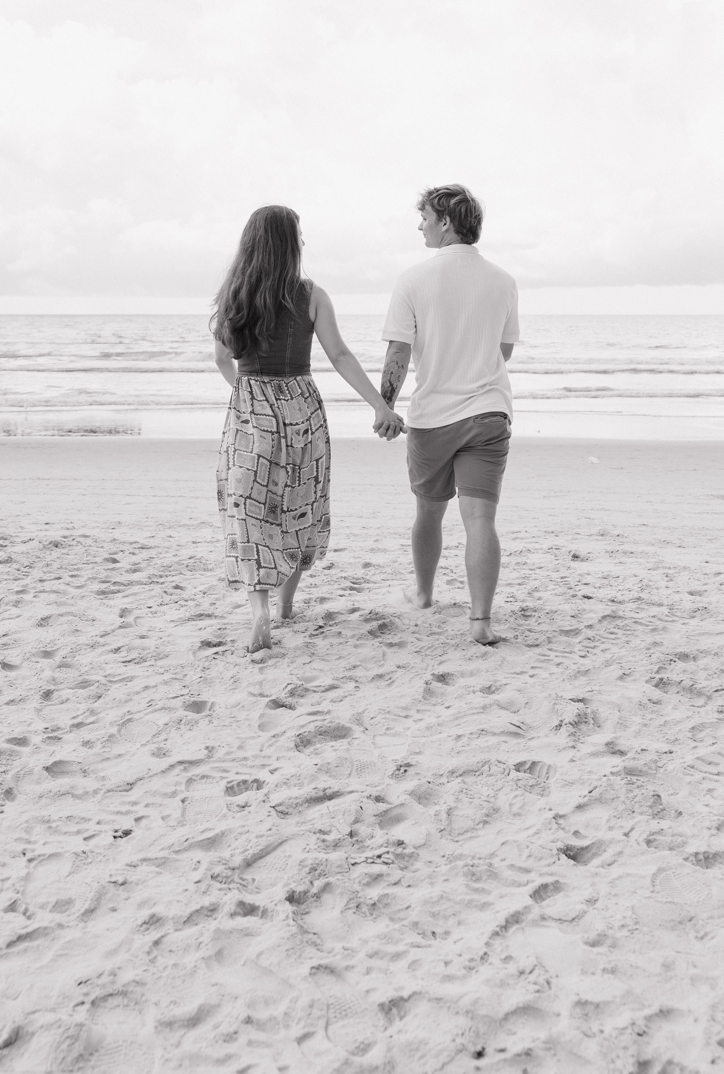 A couple walks hand in hand along a serene beach in black and white photography.